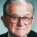 A senior man with glasses, wearing a dark suit and tie, looking directly at the camera with a neutral expression.