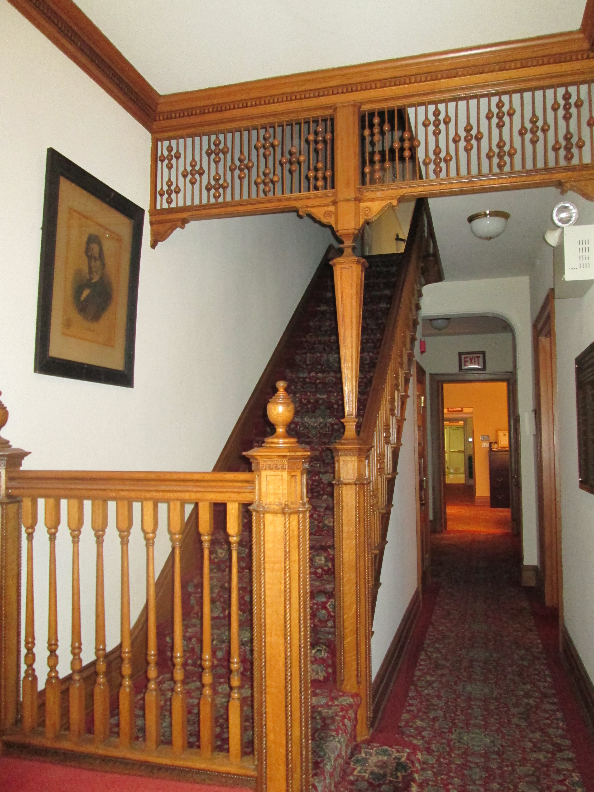 A wooden staircase with ornate railings leads up, flanked by a patterned carpet and a framed portrait on the wall.