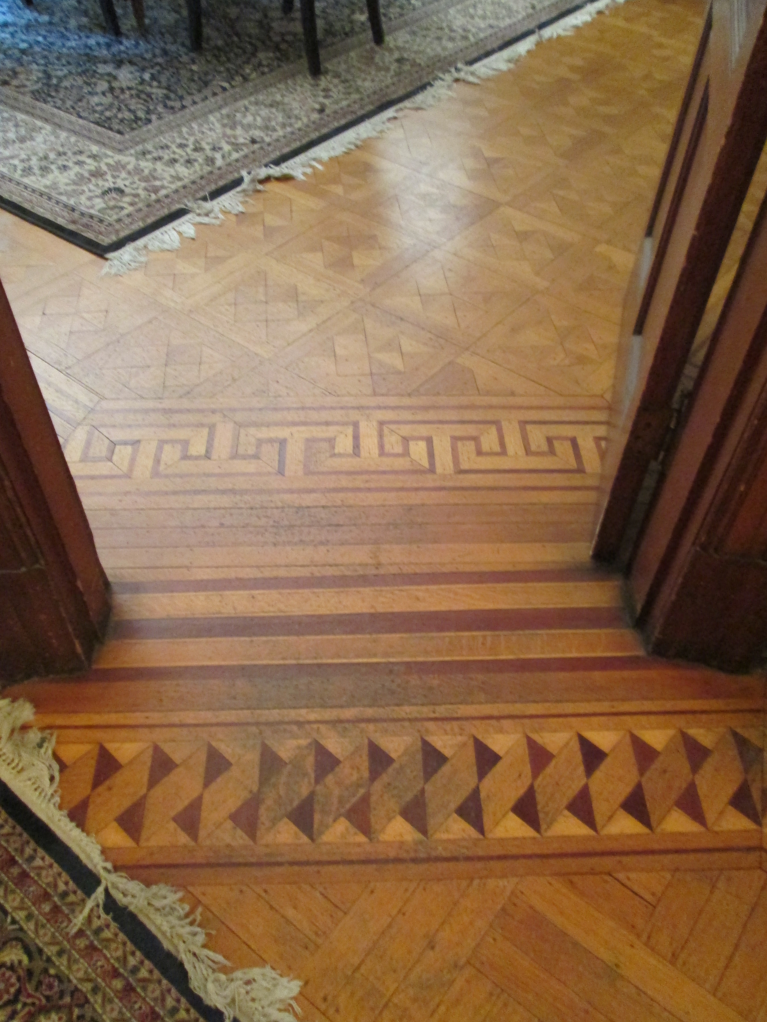 A wooden floor with intricate geometric patterns, transitioning from one design to another, with a rug visible in the background.