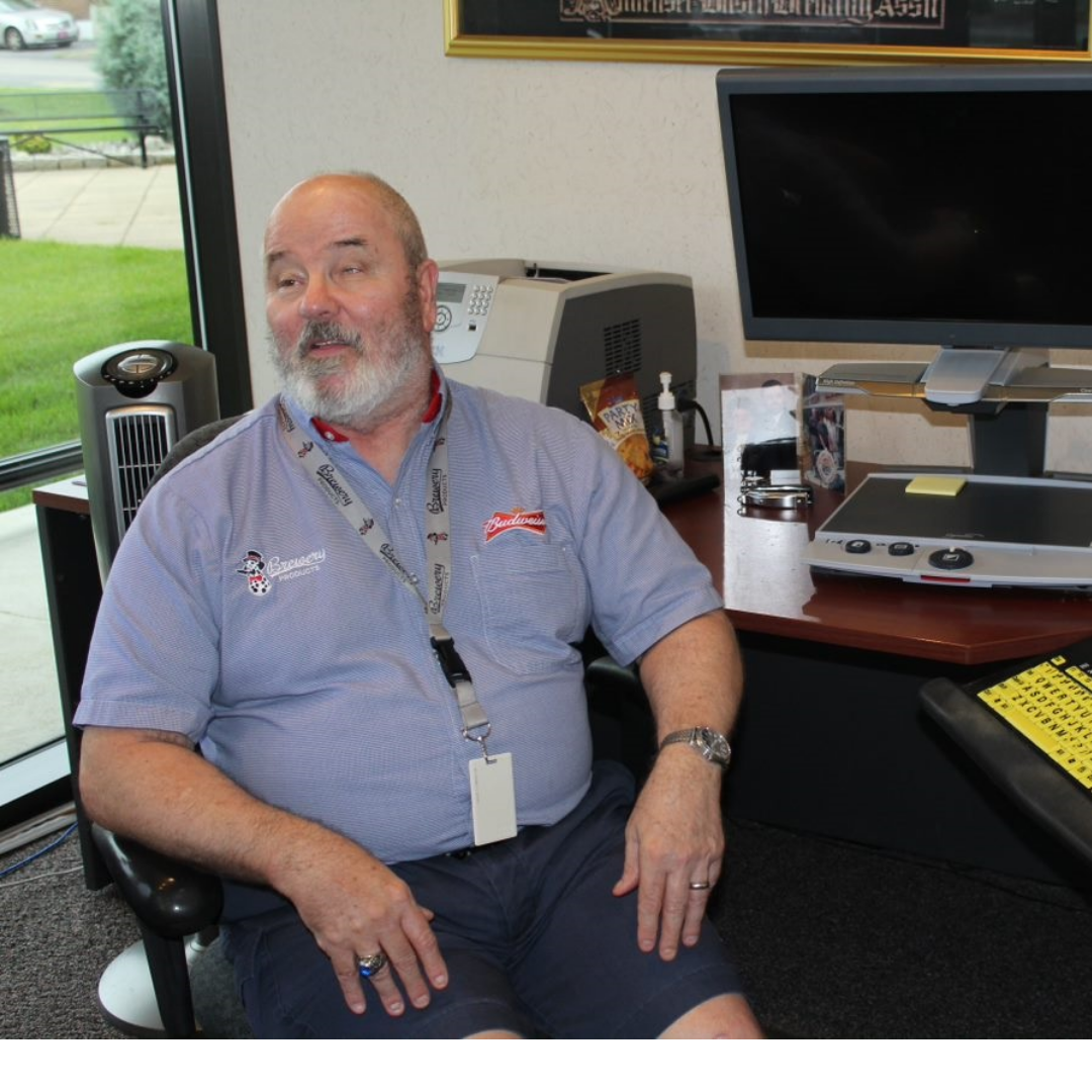 A man with a beard sits in an office chair, wearing a blue shirt and shorts, with a computer and office supplies in the background.