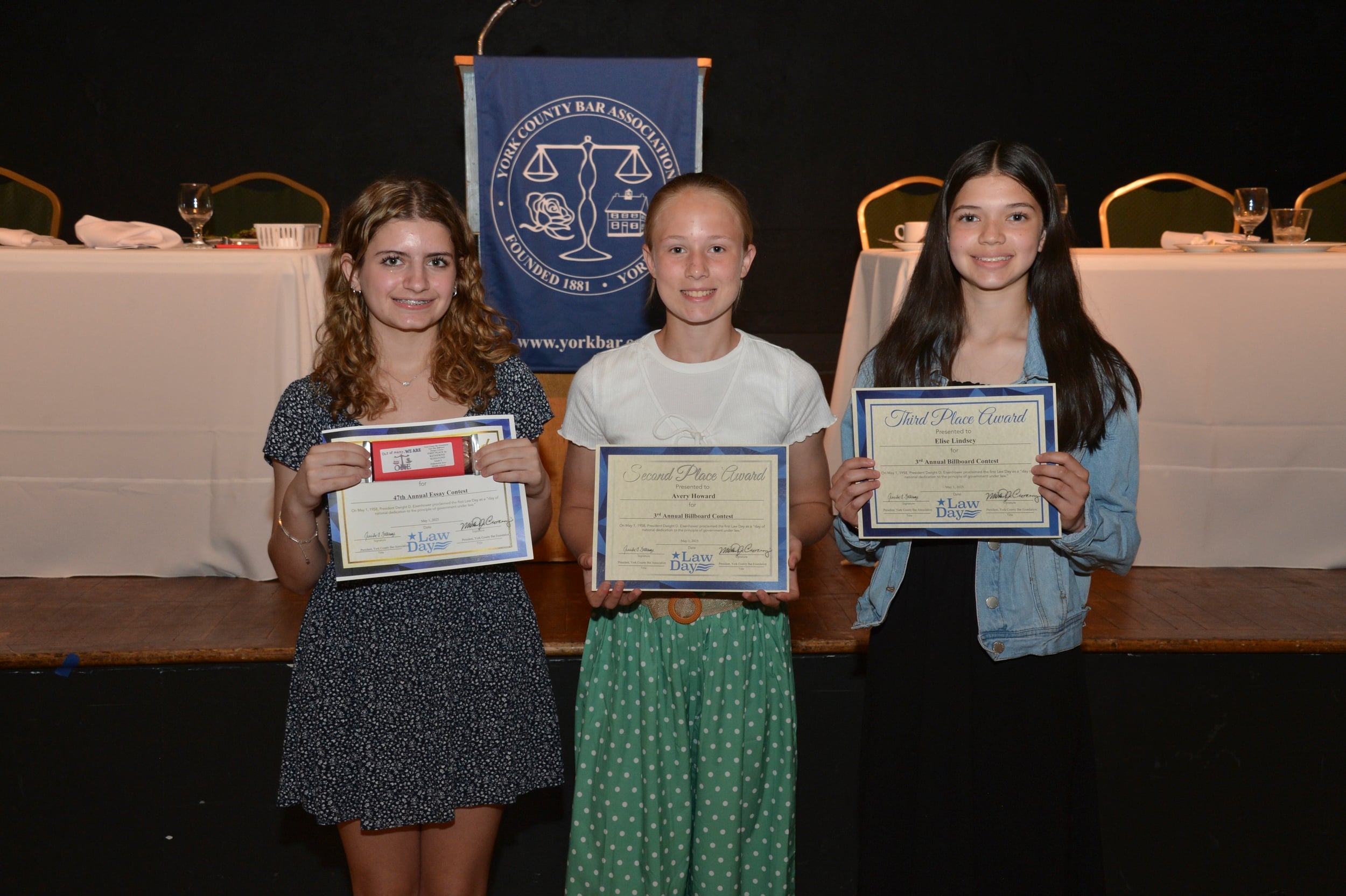 Three young girls hold awards at a ceremony, smiling. They are recognized for their achievements in an essay contest. A banner is in the background.