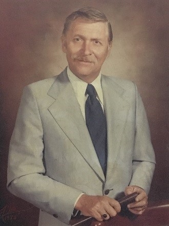 A man in a light gray suit and dark tie poses for a portrait, holding a book, with a soft, neutral background.