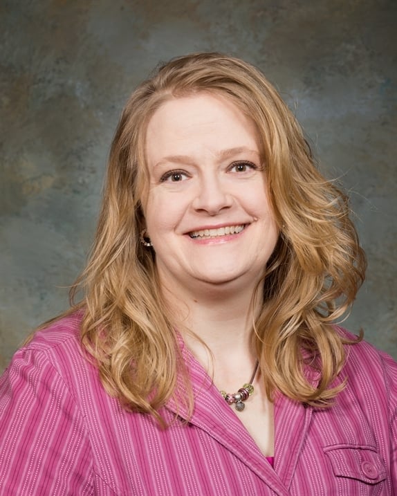 A smiling woman with long, wavy hair wearing a pink striped blouse, set against a textured background.