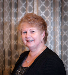 A woman with short, light brown hair smiles, wearing a black top, against a patterned curtain backdrop.